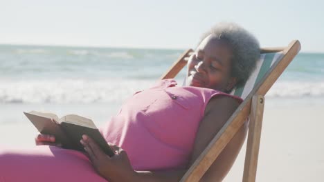 feliz mujer afroamericana mayor sentada en una silla de cubierta y leyendo un libro en la playa, en cámara lenta