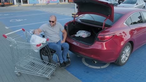 adult man with disabilities in a wheelchair puts purchases in the trunk of a car in a supermarket parking lot