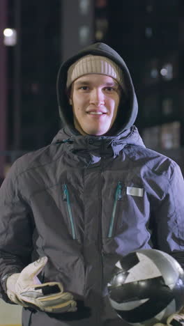 hombre sonriente con capucha alternando una pelota de fútbol en la mano mientras da un pulgar hacia arriba con la mano izquierda levantada por encima del hombro por la noche en un entorno urbano