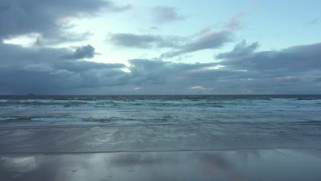 Aerial-view-over-a-person-stranding-on-a-beach,-with-hands-up-in-air,-enjoying-a-gloomy-dusk-and-waves-breaking-the-coast-of-Spain---Dolly,-drone-shot