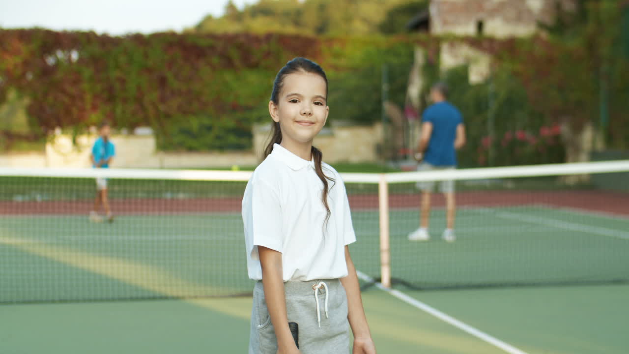 retrato de una linda niñita sosteniendo una raqueta y sonriendo alegremente a la cámara mientras estaba de pie en una cancha de tenis al aire libre
