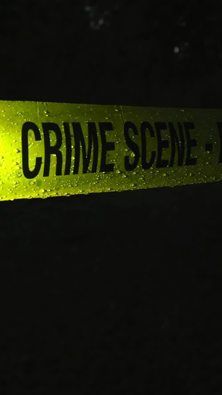 Primer plano de un vídeo de cinta policial amarilla brillante de investigación forense en la escena del crimen colocada frente a un área oscura al aire libre durante la noche mientras llueve y caen gotas de lluvia sobre ella