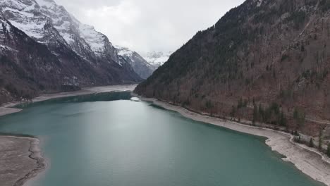 Aerial-view-of-Klöntalersee-in-Glarus,-Switzerland,-with-the-Vorderglärnisch-mountain-towering-over-the-valley-lake,-surrounded-by-snow-capped-peaks,-showcasing-the-allure-of-the-Swiss-Alps