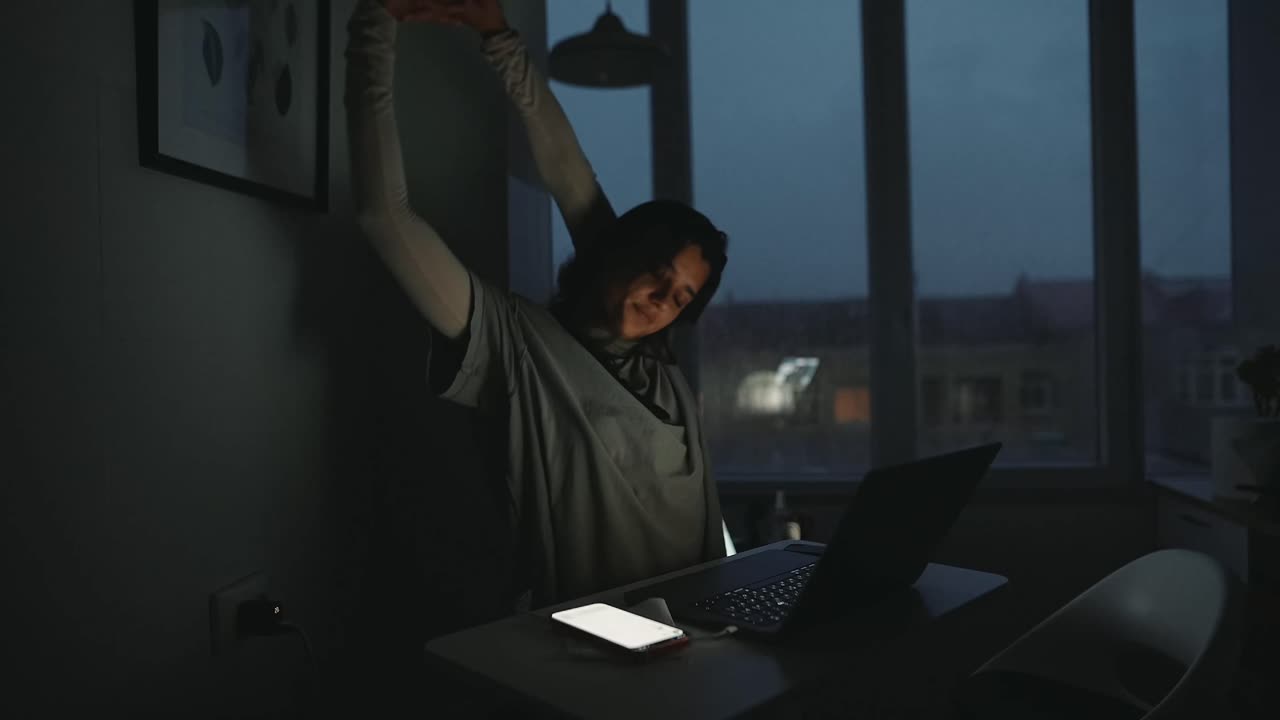 Woman stretching at desk with laptop and phone at night