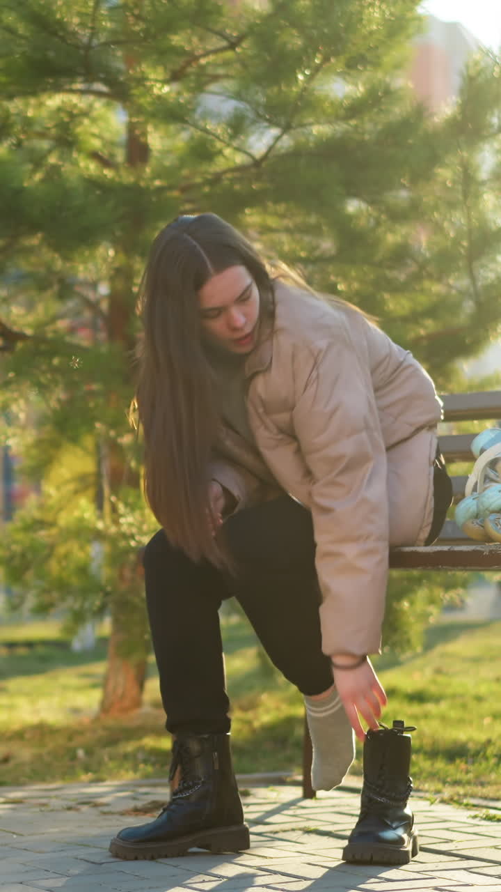 una chica con una chaqueta de melocotón y pantalones negros está sentada en un banco en un parque. se la ve quitando una bota mientras los patines descansan en el banco a su lado