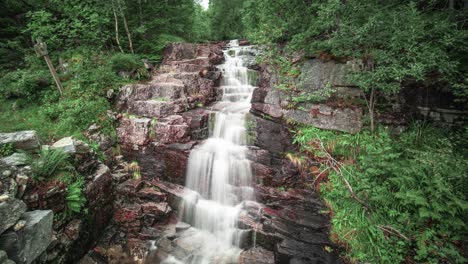 Una-Pequeña-Cascada,-Rodeada-De-Vegetación-En-El-Bosque-De-Verano,-Cae-En-Cascada-Sobre-Las-Piedras-Oscuras