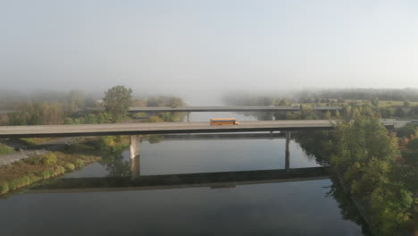 cars and school bus cross highway bridge over peaceful river with dense fog over landscape in the background