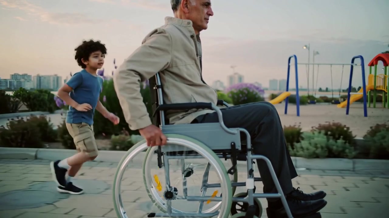 Rolling Senior propelling manual wheelchair on waterfront path, boy appearing running to keep pace