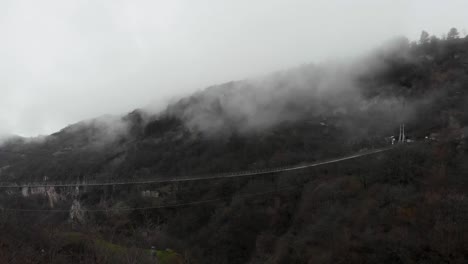 drone view of a suspension bridge for pedestrians in armenia, with dramatic clouds intermittently covering and revealing the bridge, creating a mystical atmosphere