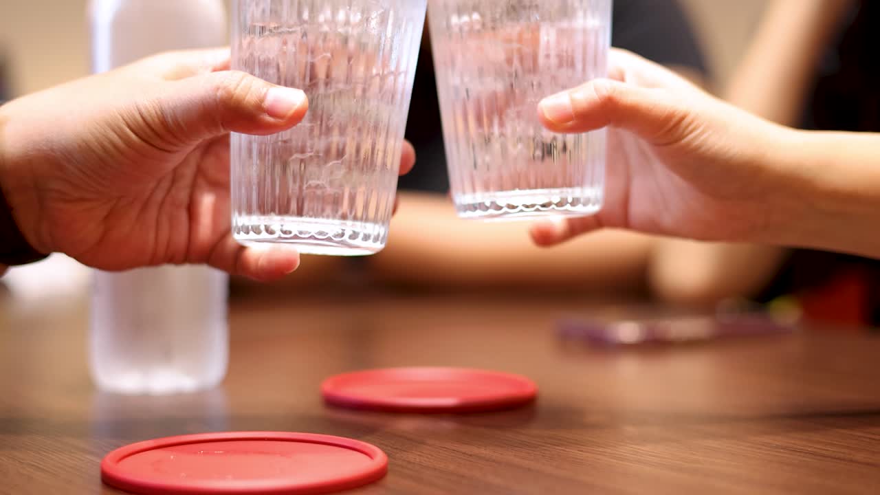 Two people raise and clink clear glasses of water over a wooden table in a warmly lit indoor setting, creating a casual, friendly atmosphere