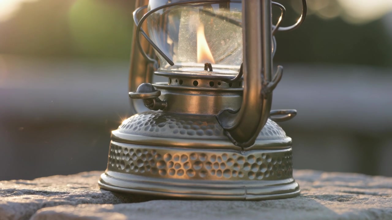 Lanterna a cherosene tremolante che mostra una fiamma inclinata su una sporgenza di pietra, con luce solare e brezza che cambiano.