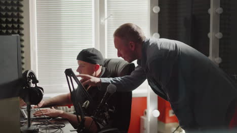 two men are in a studio room, one seated in front of a computer, likely a producer, wearing black clothes, while the other, dressed in a long sleeve shirt, leans over, explaining something intently