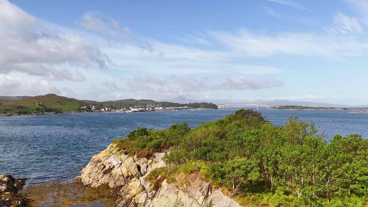 Daylight pan reveals rocky coastline, green shrubs, and calm blue waters of Loch Alsh