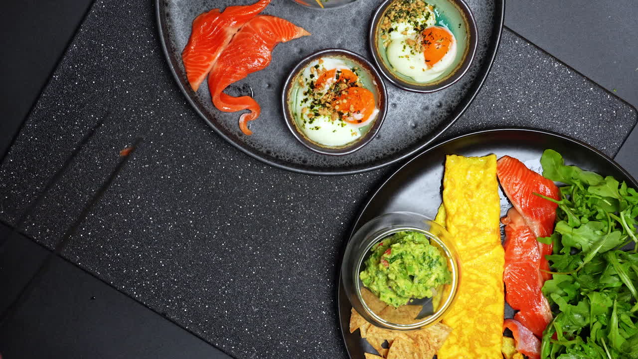 Healthy breakfast with eggs, salmon, and guacamole on a black table. Two plates of food are on a black table, showing eggs, salmon, and guacamole with chips