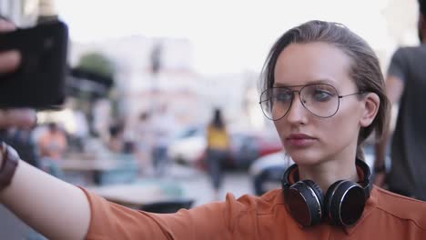 Young-woman-with-light-day-make-up-standing-on-the-street-and-taking-selfie-with-her-smartphone.-Blurred-street-background