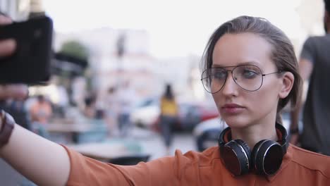 mujer joven con maquillaje de día ligero de pie en la calle y tomando selfies con su teléfono inteligente. fondo borroso de la calle