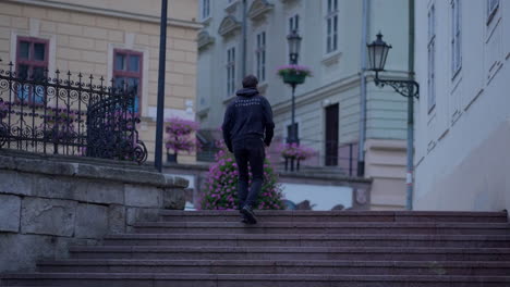 A-young-man-running-up-the-stair-in-an-old-European-city-Banska-Stiavnica,-Slovakia,-with-beautiful-historical-buildings-in-the-background-surrounded-by-purple-flowers