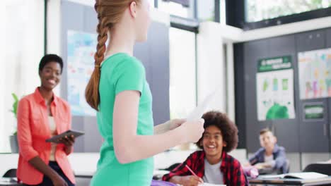 diverse female teacher and happy schoolchildren at desks reciting in school classroom