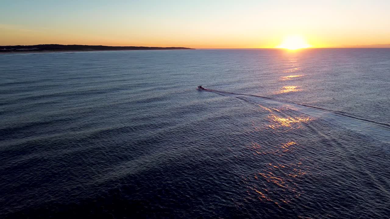 Drone aerial landscape of fishing boat cruising along ocean coastline water with waves and orange reflection from golden sunrise on horizon headland of Central Coast Australia marine tourism outdoors