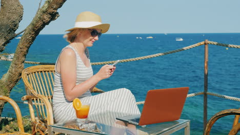 mujer turista que se relaja en un café con vista al mar disfruta de un teléfono inteligente junto a una computadora portátil