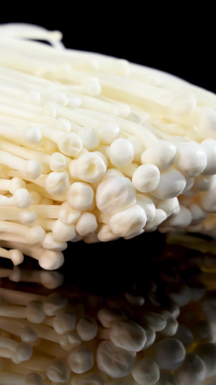Sekelompok jamur enoki berputar perlahan di permukaan hitam mengkilap, diterangi oleh pencahayaan studio lembut untuk efek makro yang bersih dan kontras tinggi.