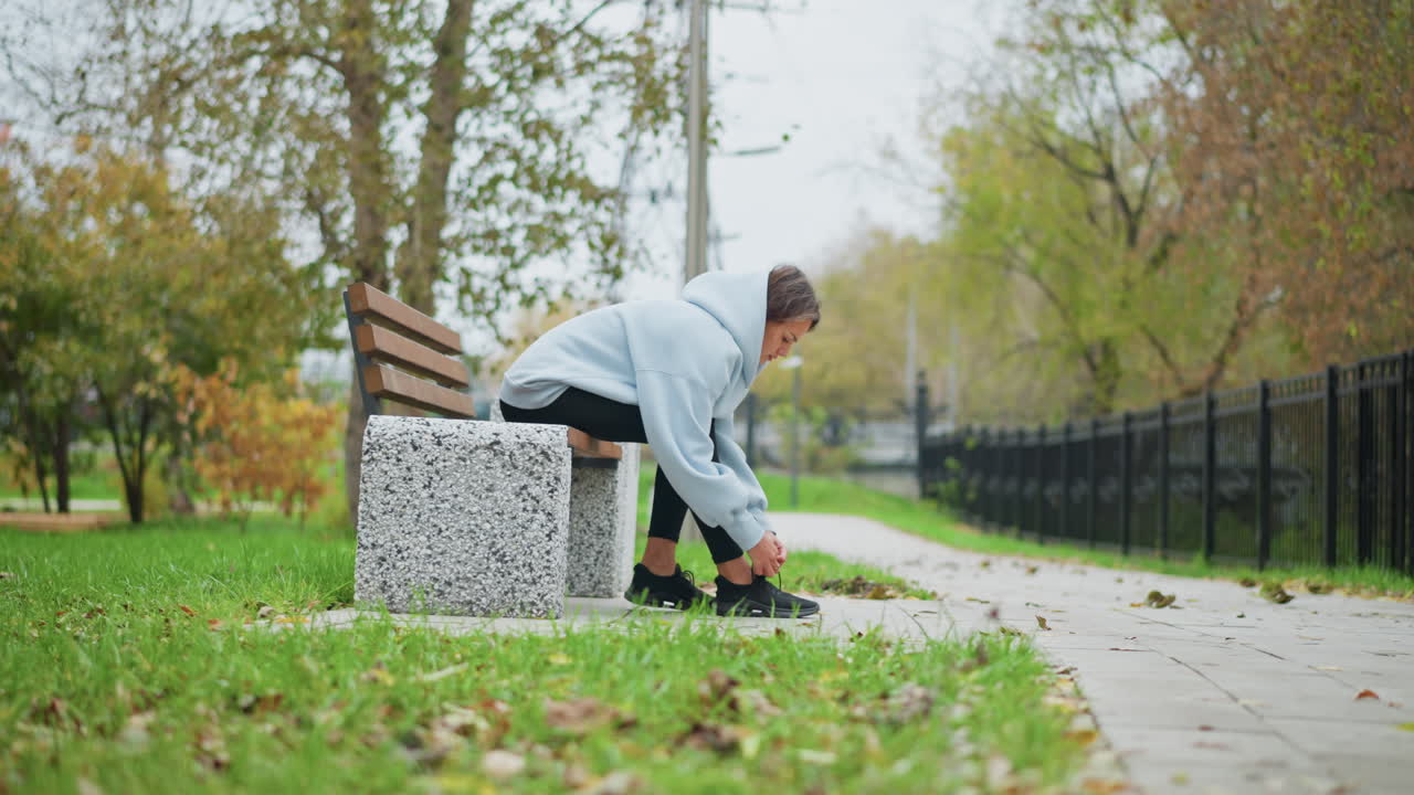 Young woman sitting on concrete bench tying her shoelace, with blurred background showing trees, iron fence, grass, and pole in an outdoor park, preparing for exercise or walk