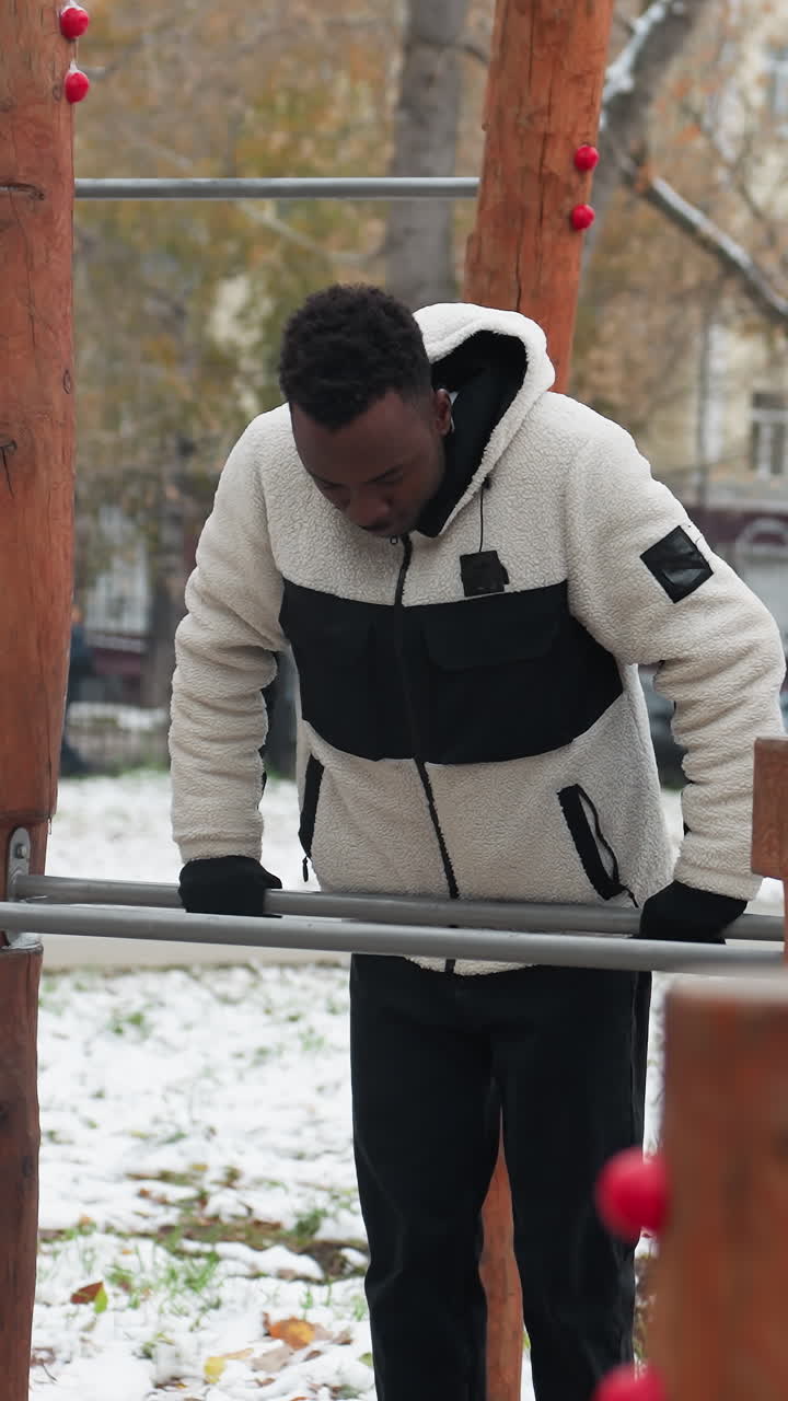 hombre negro se dedica a la rutina de fitness usando equipos de entrenamiento al aire libre durante el invierno, en medio de un entorno de ciudad ocupada con condiciones nevadas, mostrando determinación y estilo de vida urbano