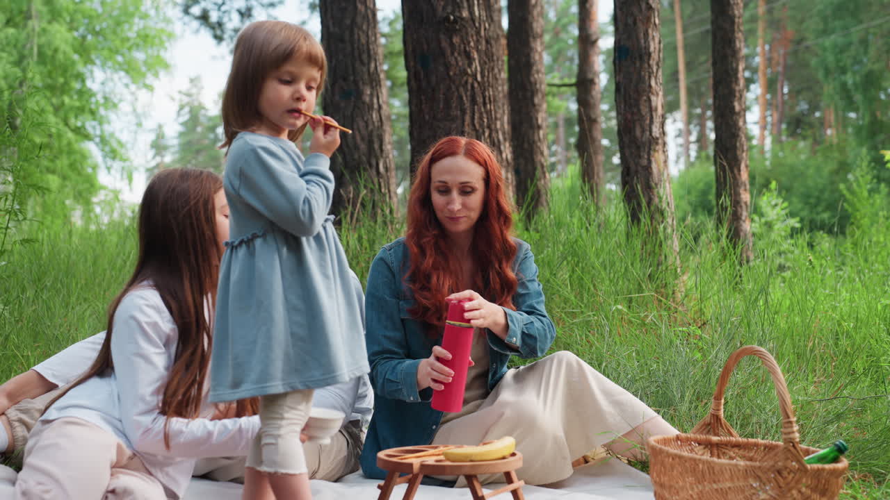 Familia feliz disfrutando de un picnic al aire libre con el padre de pie, la madre sirviendo té de un termo rojo y los niños sentados en una manta rodeados de hierba alta y árboles durante un día de verano tranquilo