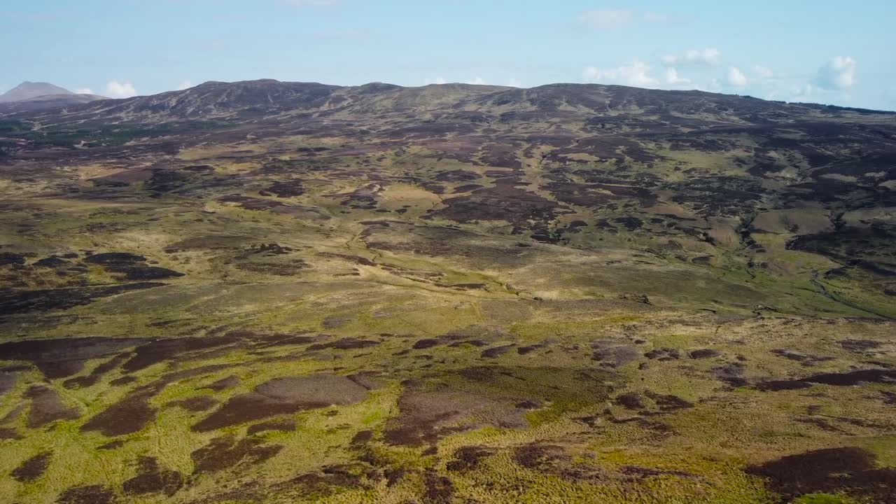 Aerial drone footage gliding and flying over Scotland gorgeous brown and green grassy and moss covered terrain landscape with a large hill or a mountain in the background. Sky is blue with some clouds
