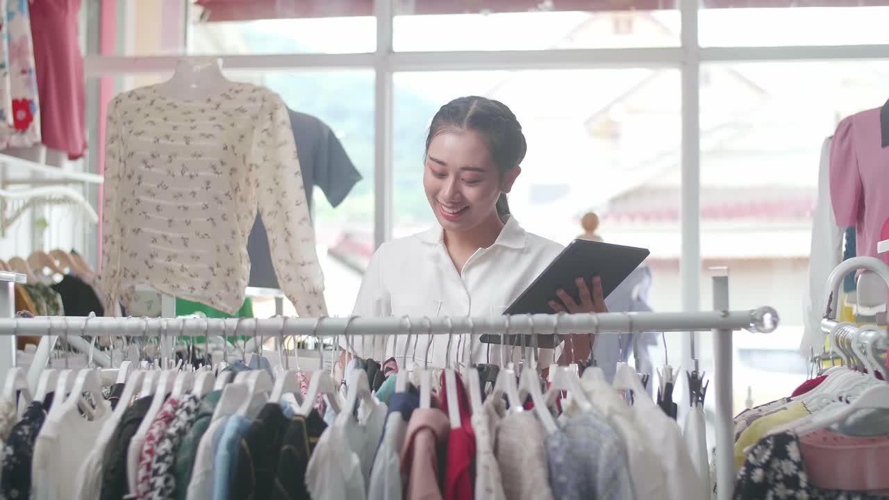 의류 가게의 아시아 여성 소유자는 디지털 태블릿을 사용하여 레일에 있는 재고를 확인하는 것을 즐긴다