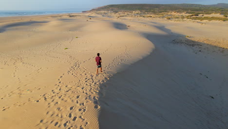 Vista-Aérea-De-Un-Joven-Caminando-Sobre-Una-Duna-Durante-La-Puesta-De-Sol-En-La-Playa-De-Frontera,-Portugal