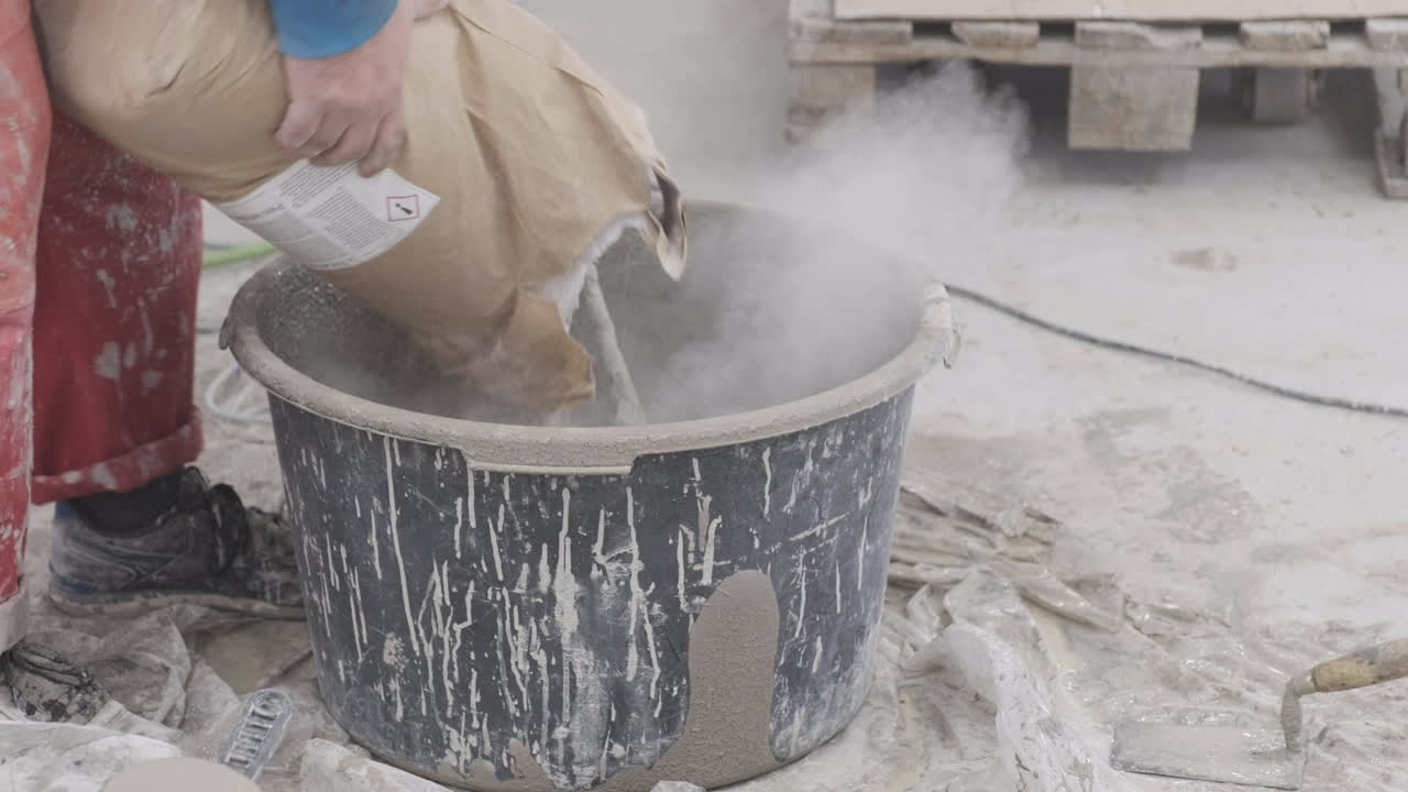 Pouring powder from a bag into a bucket The worker is wearing overalls It appears to be a construction setting There is powder in the air