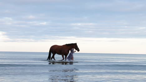 Una-Hermosa-Chica-Con-El-Pelo-Largo-Y-Un-Vestido-Azul-Pasea-A-Su-Caballo-Por-El-Agua-Durante-La-Noche-En-Donabate,-Irlanda