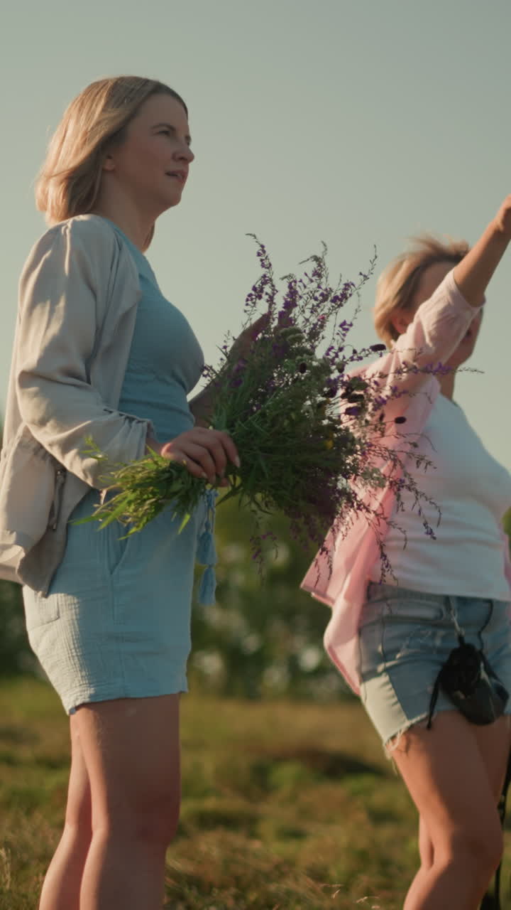 vista lateral de la familia de pie en tierras de cultivo montañosas bajo un cielo despejado mientras la madre señala algo interesante en la distancia a su hija, mientras que otra mujer está de pie cerca sosteniendo un ramo de flores silvestres
