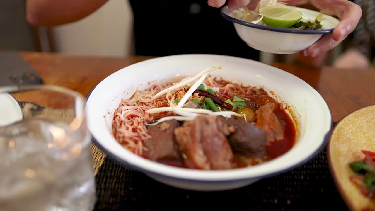A hand adds fresh bean sprouts to a bowl of rice vermicelli with Northern Thai curry sauce in a well-lit Bangkok restaurant, shot in close-up