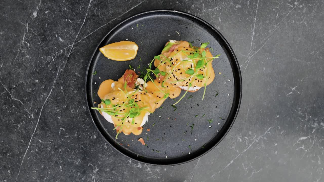 Top view of a gourmet dish with poached eggs, sauce, and fresh herbs on a black plate with lemon wedge, beautifully styled on a dark marble surface, spinning shot