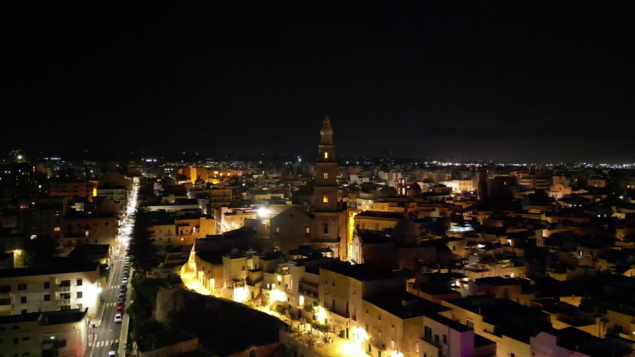 Aerial view of Monopoli at night with illuminated streets and the cathedral tower standing out in the city lights.