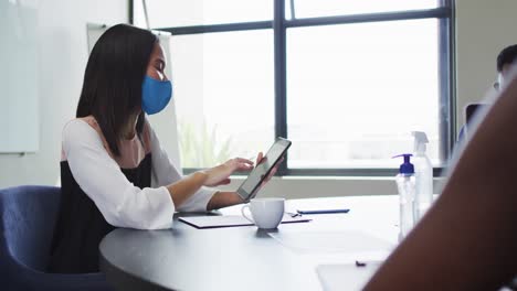 Asiatische-Frau-Mit-Gesichtsmaske-Und-Digitalem-Tablet,-Während-Sie-Auf-Ihrem-Schreibtisch-Im-Modernen-Büro-Sitzt