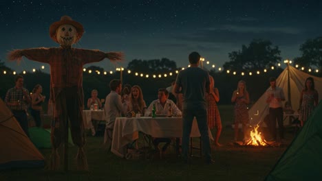 a festive outdoor gathering under starry skies featuring a scarecrow with guests enjoying food, drinks, and warmth by the campfire amid string lights and tents