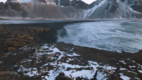 Aerial-view-tilting-over-the-snowy-coast-of-Stokksnes,-revealing-the-Vestrahorn-mountain,-sunset-in-Iceland