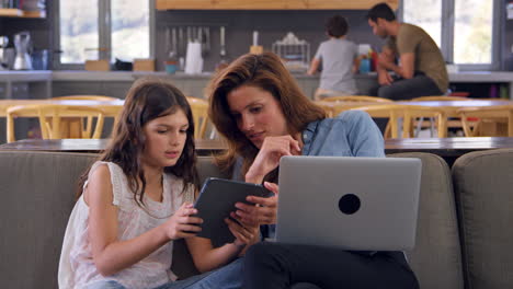 mother and daughter sitting on lounge sofa using digital devices