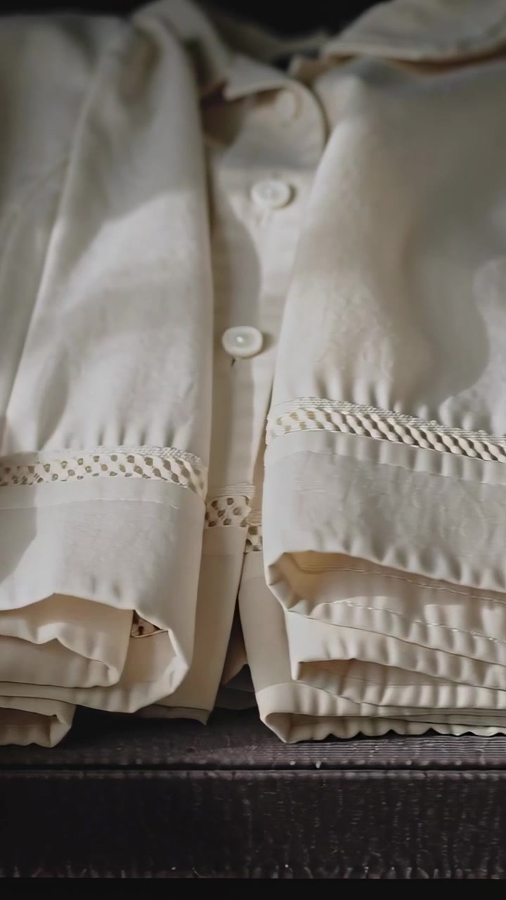 Video verticale: Mani che sollevano e rivelano camicie color crema in una valigia, lisciando le rifiniture intrecciate per preparare il bagaglio