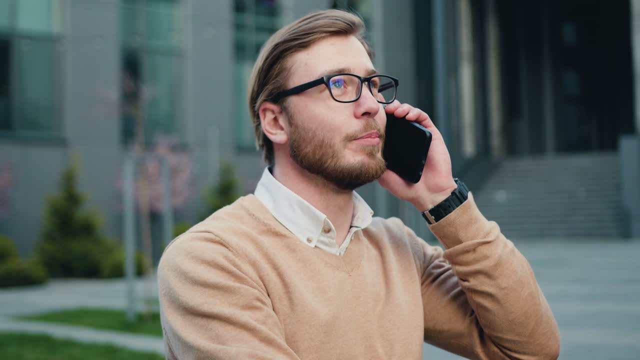 Close up of successful bearded man in glasses talking cellphone in the city financial district. Outdoors