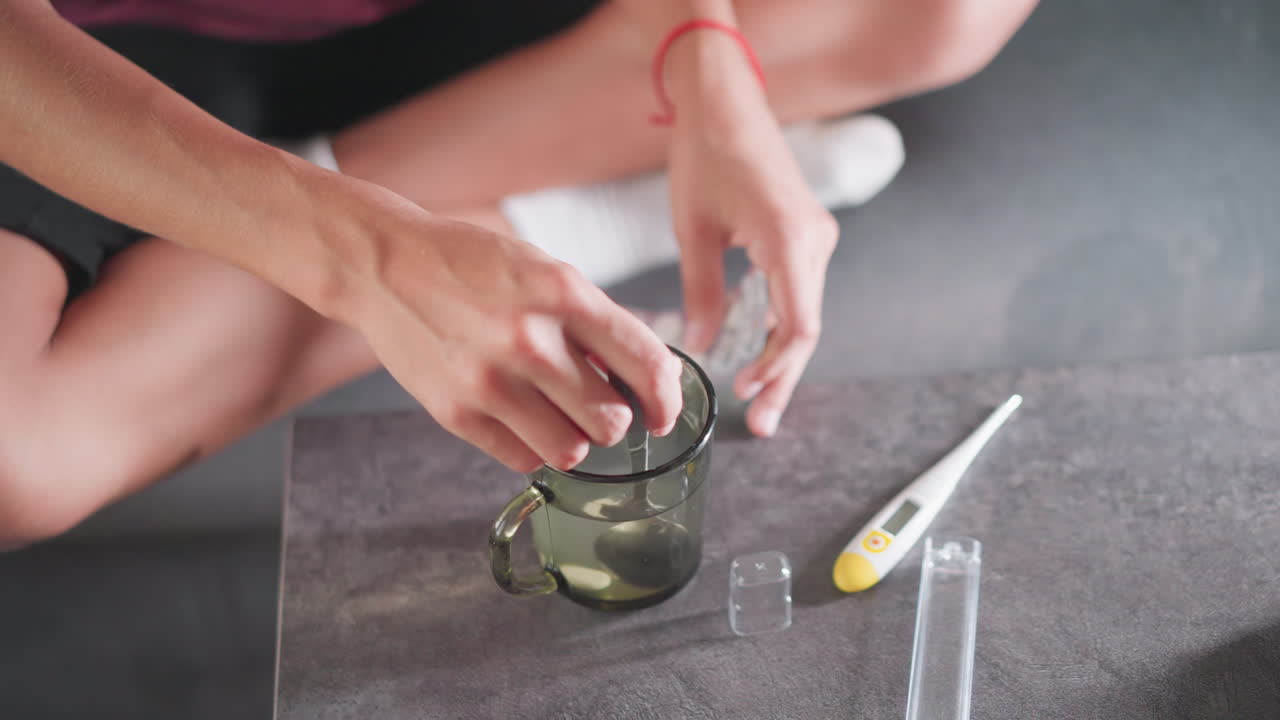 Close up of individual seated on floor stirring drug in glass of water using spoon while surrounded by medical tools including thermometer, pill packs, and clear plastic covers