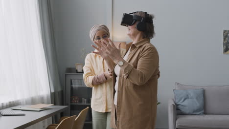 Lächelnde-Arabische-Frau-Mittleren-Alters,-Die-Einer-Glücklichen-älteren-Dame-Hilft,-Zu-Hause-Eine-Virtual-reality-headset-brille-Zu-Verwenden