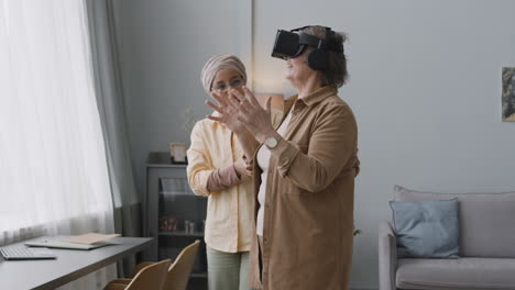 smiling middle aged arabic woman helping a happy senior lady to use virtual reality headset glasses at home