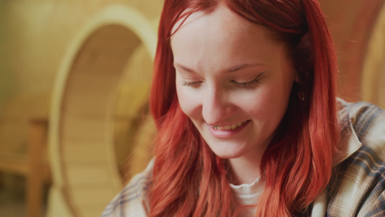 caucasian woman journaling in cozy cafe, soft warm lighting, wooden interior, flannel shirt and red hair, smiling while writing in notebook, laptop on table, barista background, study and creative