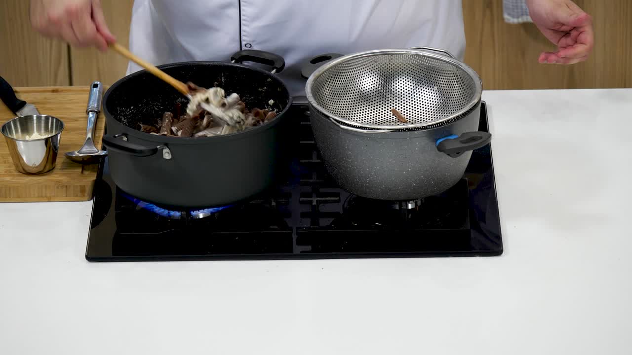 A chef skillfully drains cooked pasta into a colander above a pot on a gas stove while preparing a flavorful dish in a sleek kitchen setup. The activity highlights culinary techniques