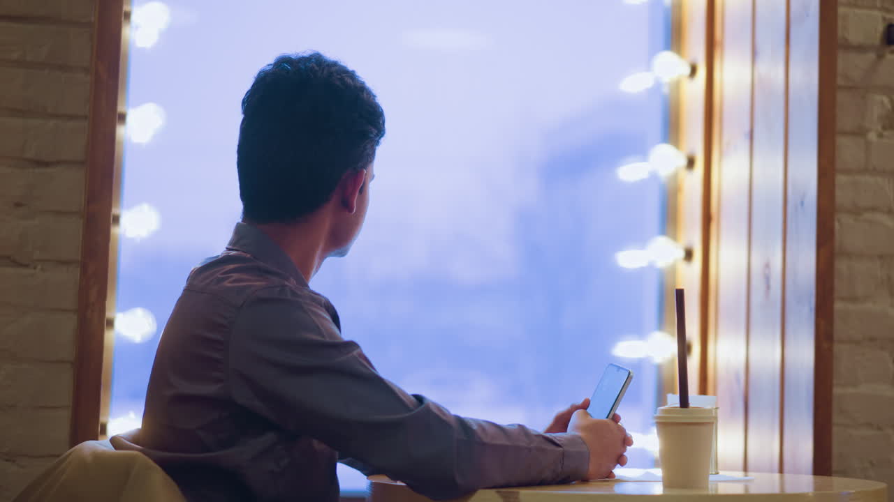 Man sitting alone in cozy cafe holding phone, looking out large window with soft lights glowing, lost in thought, coffee cup on table, wintery urban backdrop visible through frosted glass