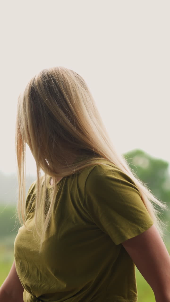 금발 머리카락을 가진 예쁜 젊은 여성은 비가 오는 날 휴양지에서 흐릿한 산의 실루을 대비하여 울창한 을 둘러보고 서서히 움직입니다.